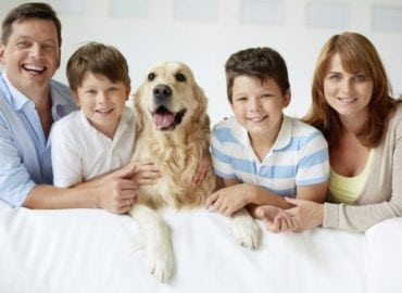 A happy family consisting of a mother, father, two boys, and their golden retriever, all smiling and sitting together on a white sofa.