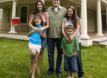 A happy family of five, with two adults and three children, standing in front of their new home with a "Sold" sign visible. They are smiling in a grassy front yard.