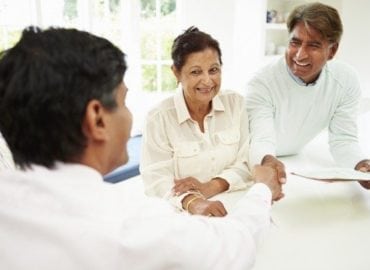 A senior couple happily consulting with a male professional at a desk in a bright, sunlit room. The woman is smiling broadly while shaking hands with the professional.