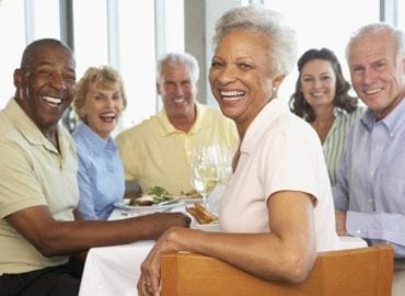 A group of five elderly friends, three men and two women, sitting around a table in a restaurant, smiling and enjoying a meal together.