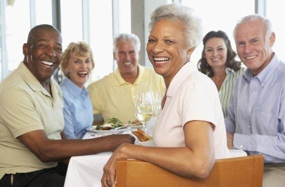 A group of five elderly friends, three men and two women, sitting around a table in a restaurant, smiling and enjoying a meal together.