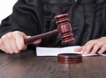 A judge in a black robe is about to strike a wooden gavel on a sounding block, with a document visible on the desk in front of them.