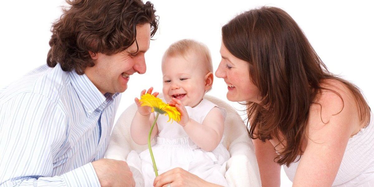 A joyful family with a mother, father, and baby interacting with a yellow flower, all smiling and dressed in light, casual clothing against a white background.