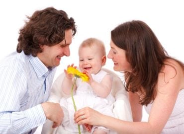 A joyful family with a mother, father, and baby interacting with a yellow flower, all smiling and dressed in light, casual clothing against a white background.