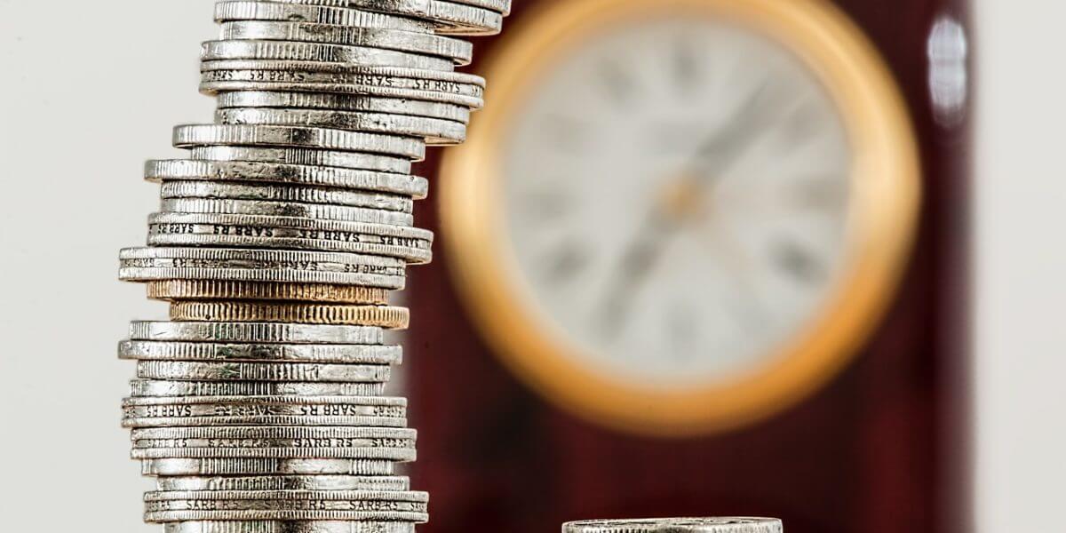 A close-up image of a stack of silver coins with a blurred clock in the background, suggesting themes of finance and time management.
