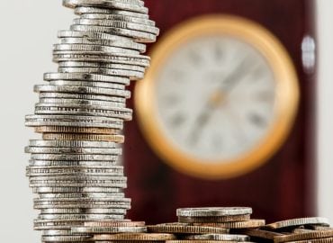 A close-up image of a stack of silver coins with a blurred clock in the background, suggesting themes of finance and time management.