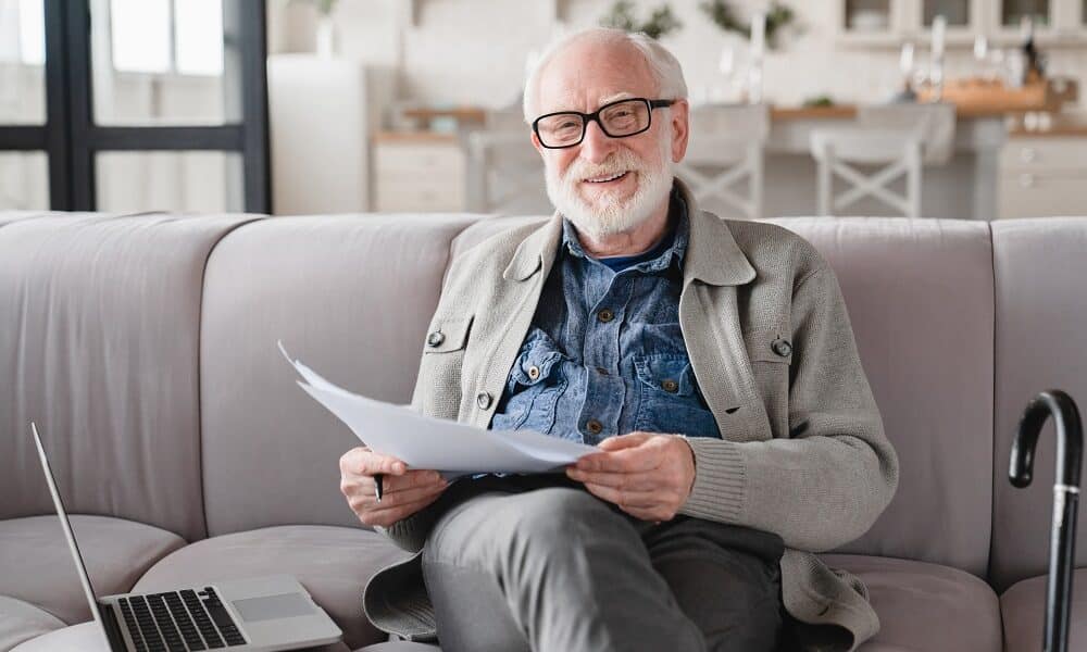 Old elderly senior caucasian man grandfather looking at camera checking pension documents, loan, e-banking, doing paperwork with laptop, mortgage, paying domestic bills at home