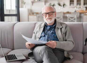 Old elderly senior caucasian man grandfather looking at camera checking pension documents, loan, e-banking, doing paperwork with laptop, mortgage, paying domestic bills at home