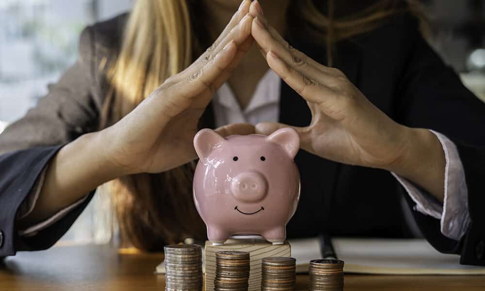 A person in a suit encircles their hands protectively over a pink piggy bank. Multiple stacks of coins are arranged in front of the piggy bank on a wooden table, symbolizing savings, financial security, and asset protection.
