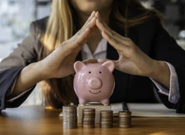 A person in a suit encircles their hands protectively over a pink piggy bank. Multiple stacks of coins are arranged in front of the piggy bank on a wooden table, symbolizing savings, financial security, and asset protection.