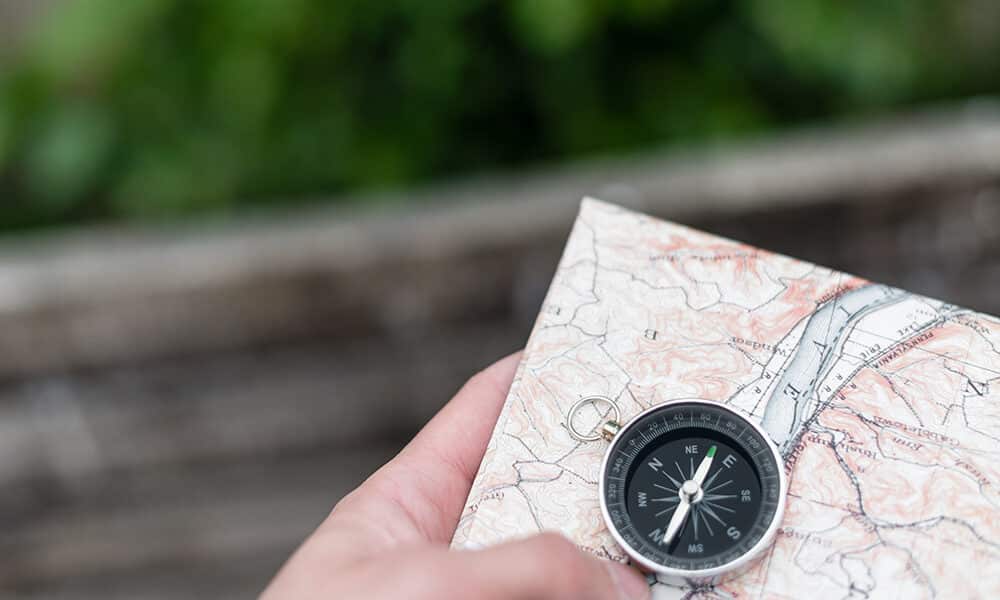 A hand holding a partially folded map with a compass placed on top of it, symbolizing navigating the challenges of co-parenting. The background is blurred, showing an outdoor setting with green foliage.