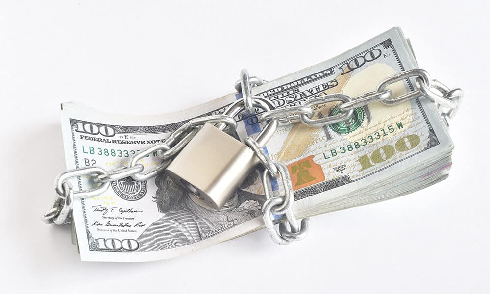 A stack of 100-dollar bills is wrapped in a metal chain with a padlock securing it, symbolizing how proactive planning can shield your assets from potential threats. The arrangement is set against a plain white background.
