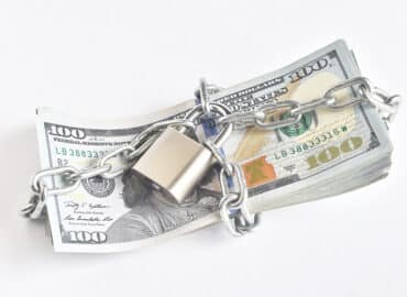 A stack of 100-dollar bills is wrapped in a metal chain with a padlock securing it, symbolizing how proactive planning can shield your assets from potential threats. The arrangement is set against a plain white background.