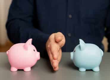 A person is placing their hand vertically between a pink piggy bank on the left and a blue piggy bank on the right, symbolizing the division of separate assets in a divorce. The individual, wearing a dark shirt, clearly signifies separation or organization of belongings during this process.
