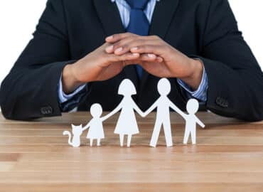 A businessman in a suit places his hands protectively over paper cutouts of a family on a wooden table. The cutouts include a father, mother, two children, and a cat, symbolizing the concept of family protection and life insurance.