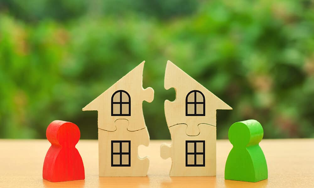 Two wooden puzzle pieces shaped like houses are connected in the middle, representing marital property. They have windows drawn on them. On each side of the houses, there are human-shaped wooden figures, one red and one green, standing on a wooden surface against a blurred green background.