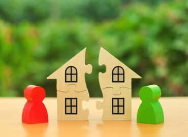 Two wooden puzzle pieces shaped like houses are connected in the middle, representing marital property. They have windows drawn on them. On each side of the houses, there are human-shaped wooden figures, one red and one green, standing on a wooden surface against a blurred green background.