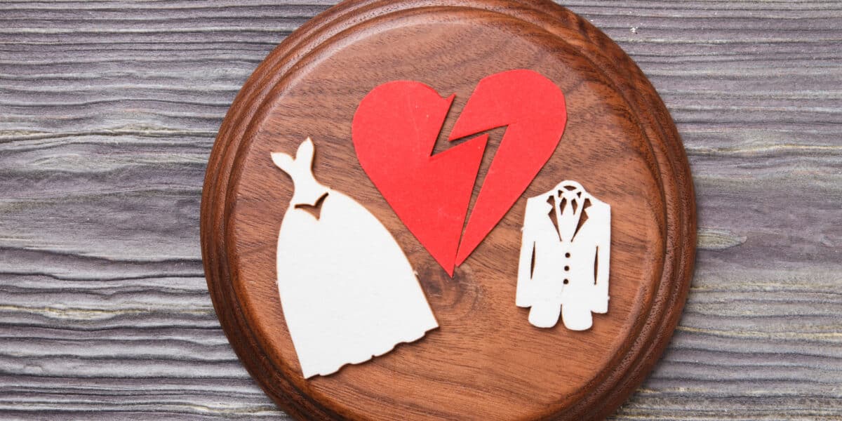 A broken red heart is set against a wooden background. On either side of the heart, there are two wooden cutouts: a wedding dress on the left and a suit on the right, symbolizing divorce or annulment.