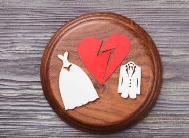 A broken red heart is set against a wooden background. On either side of the heart, there are two wooden cutouts: a wedding dress on the left and a suit on the right, symbolizing divorce or annulment.