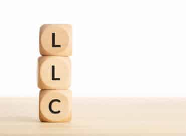 Three wooden blocks stacked vertically, each with a black letter, starting from the top: "L", "L", "C" against a plain white background. The blocks rest on a light wooden surface, symbolizing how LLCs can protect your assets with simplicity and strength.