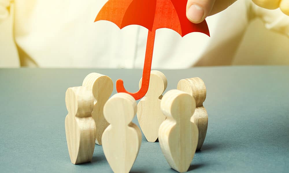 A hand holds a small red umbrella over a group of wooden human figures. The background is plain, emphasizing the concept of insurance protection and safety for the group.