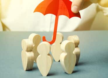 A hand holds a small red umbrella over a group of wooden human figures. The background is plain, emphasizing the concept of insurance protection and safety for the group.