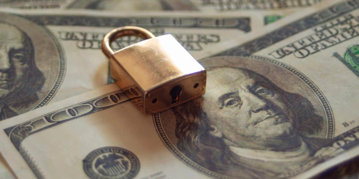 A brass padlock rests on a spread of hundred-dollar bills, symbolizing financial protection with a secure shield. The image highlights the concept of asset protection trusts and security against creditors, emphasizing the importance of safeguarding one's finances.