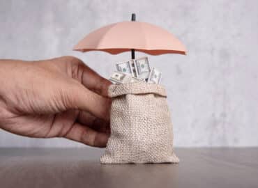 Hand holding umbrella protecting bag of money.