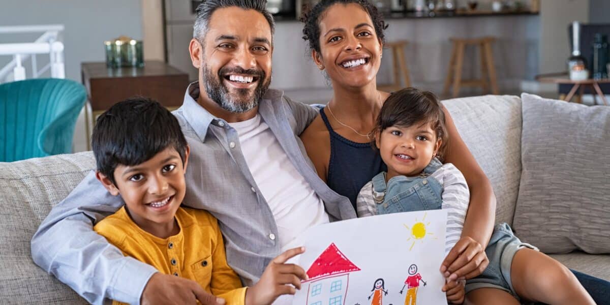 Proud parents showing family painting of son and daughter sitting on sofa at home. Smiling african mother and middle eastern father with two children looking at camera. Little boy and cute girl with parents showing handmade painting of a happy united family in a new home.