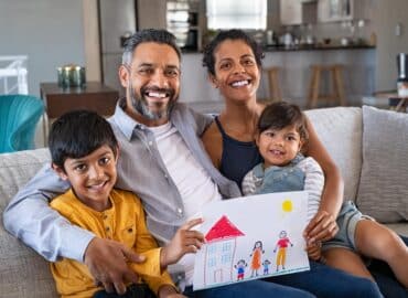 Proud parents showing family painting of son and daughter sitting on sofa at home. Smiling african mother and middle eastern father with two children looking at camera. Little boy and cute girl with parents showing handmade painting of a happy united family in a new home.
